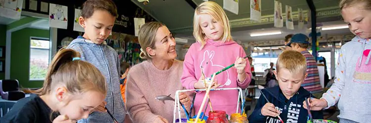 Teacher in a vibrant classroom working with primary school aged children painting at a shared table.